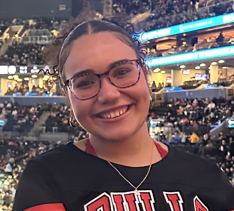 A woman with dark curly hair and glasses smiles for a photo at a crowded indoor arena. They wear a black Chicago Bulls shirt and a necklace, with multiple levels of stadium seating filled with spectators and blue illuminated signage visible in the background.