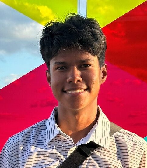 A man in a striped shirt poses in front of colorful geometric panels in yellow, red, and blue tones, with a city skyline and water visible in the background from an elevated viewpoint.