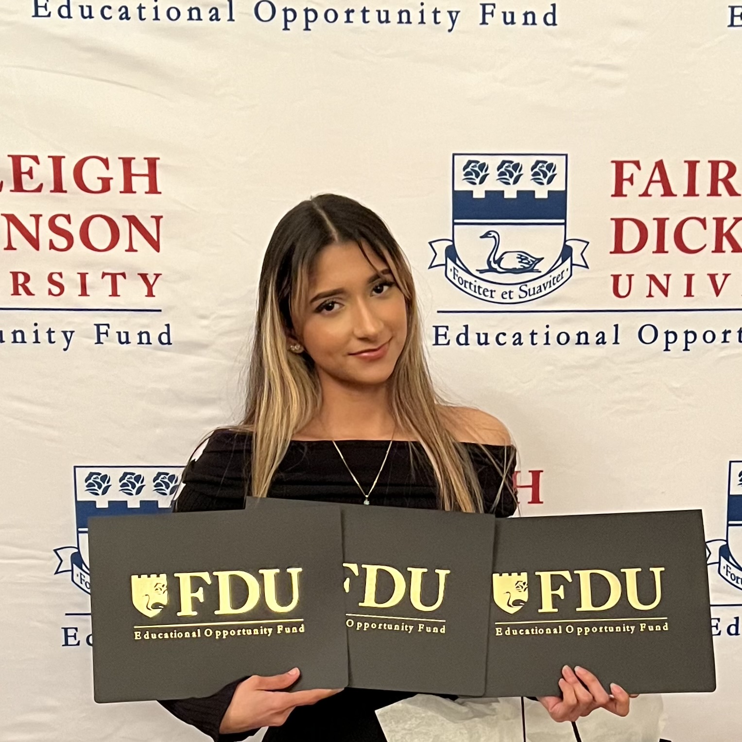 Student at an FDU Educational Opportunity Fund event holds three program folders while standing in front of a university banner featuring the school's swan crest logo.