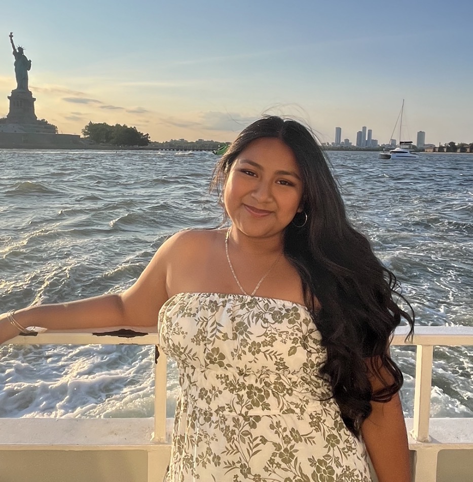 Woman in a floral dress poses on a boat with the Statue of Liberty and NYC skyline visible in the background across the water during golden hour.
