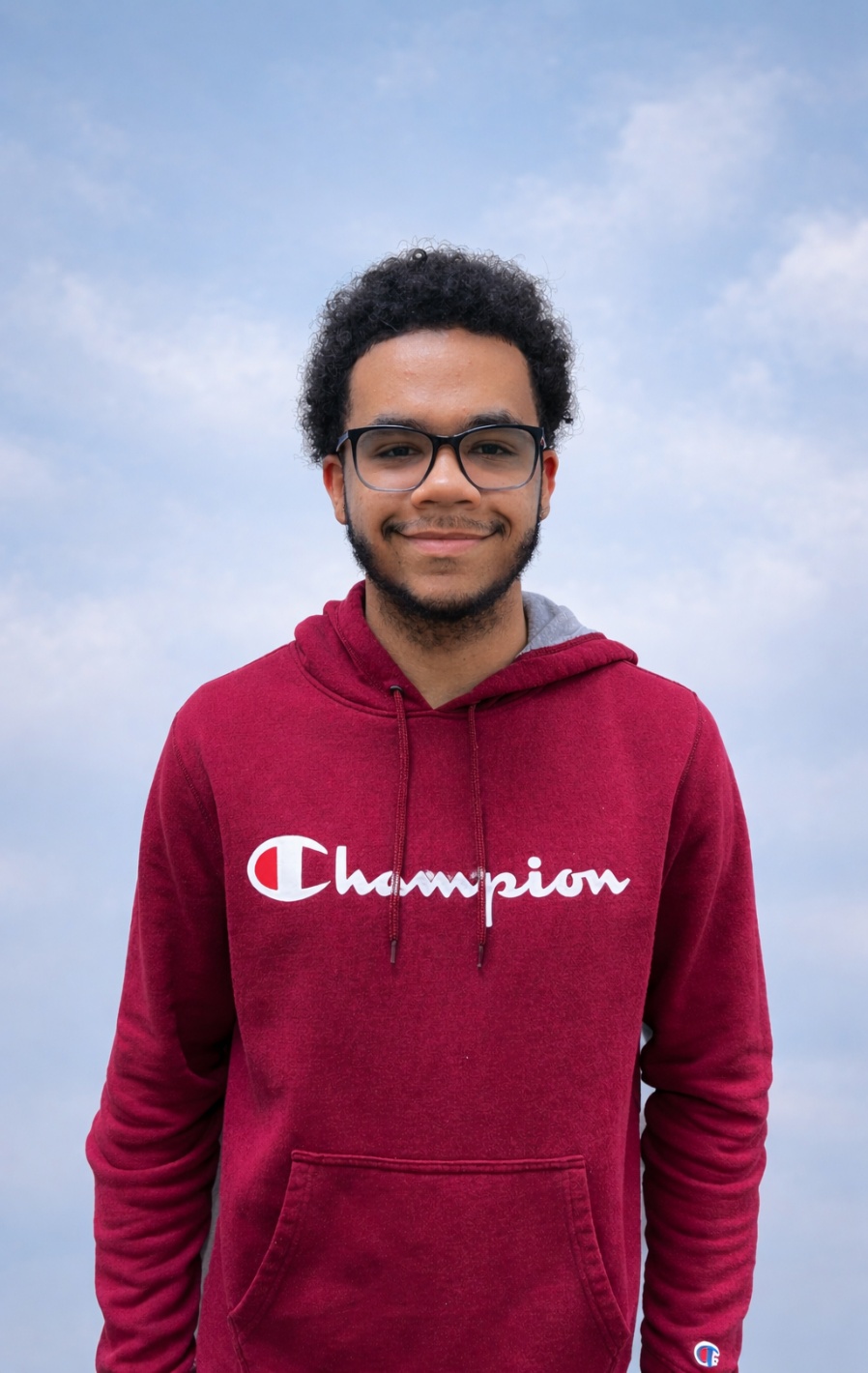 A man wearing glasses and a maroon Champion hoodie stands outdoors against a blue sky with clouds, photographed from below.