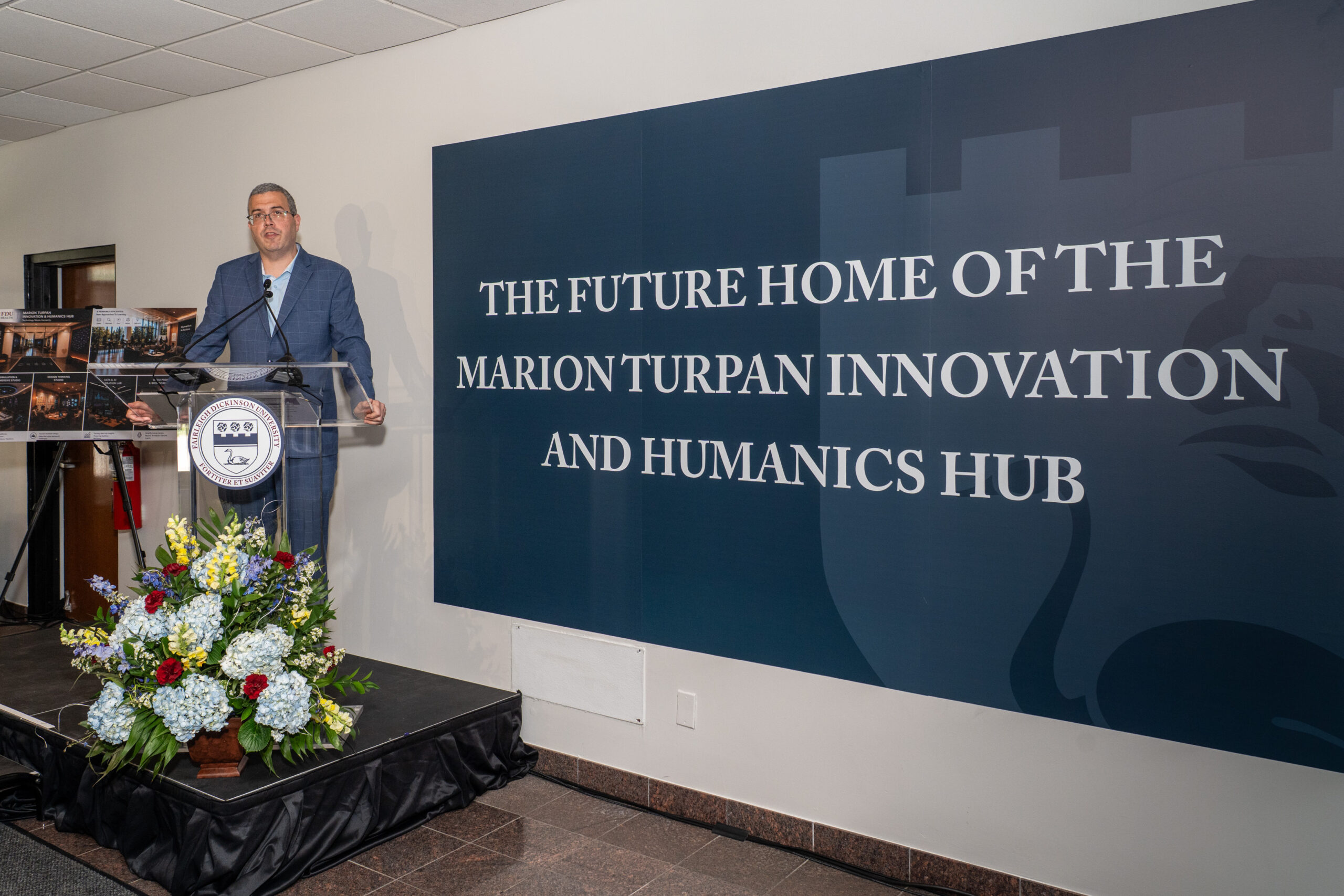 A speaker at a podium with the Fairleigh Dickinson University seal addresses an audience at an event announcing the future home of the Marion Turpan Innovation and Humanics Hub.