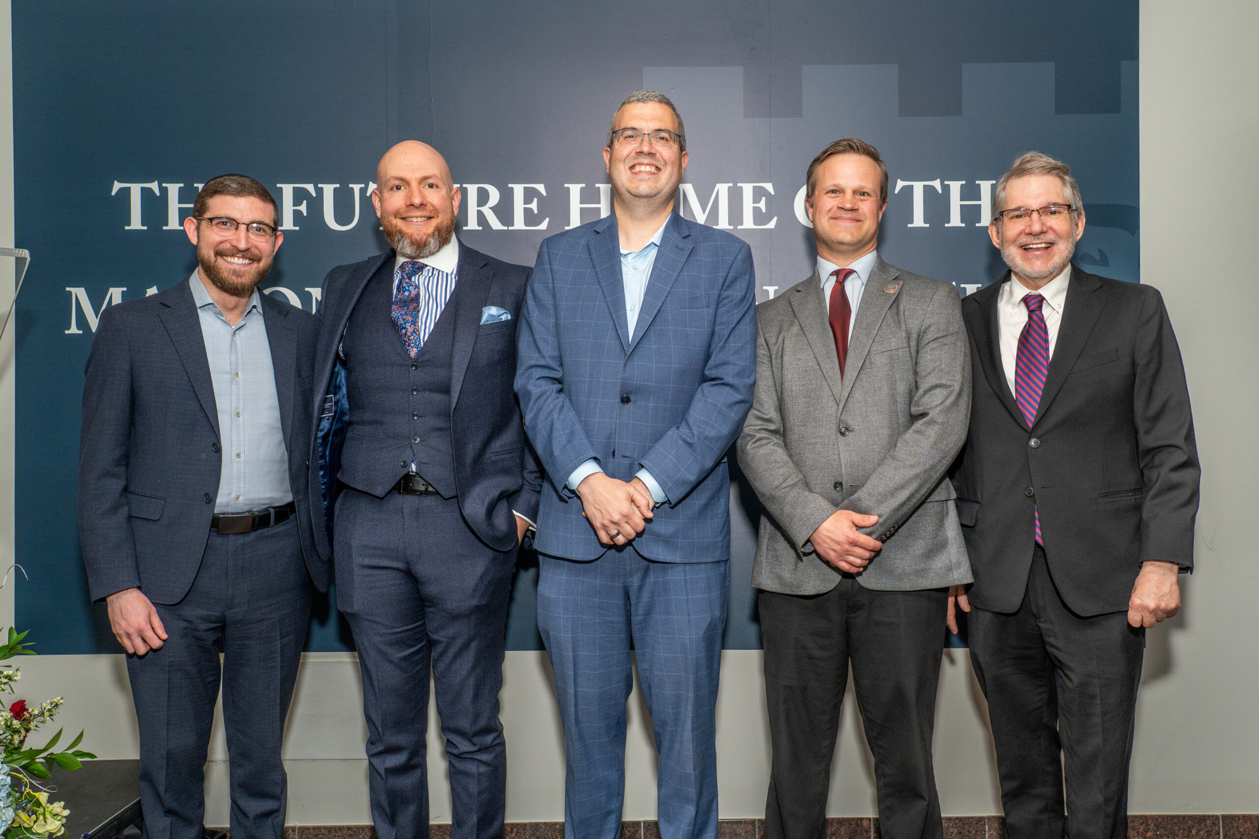 five men in business attire smile for a group photo in front of the Marion Turpan Innovation and Humanics Hub banner at a Fairleigh Dickinson University event.