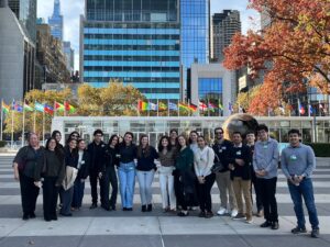 A large group poses outdoors on a plaza with a golden globe sculpture behind them. International flags line the area in front of modern glass skyscrapers. Trees with orange and yellow fall foliage frame the scene, and the group wears casual autumn clothing with name tags.