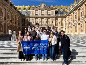 Students and adults stand on marble steps holding a blue "Fairleigh Dickinson University" banner. Behind them is an ornate palace courtyard with red brick and stone architecture, gold decorative elements, statues, and a checkered floor pattern under a blue sky.