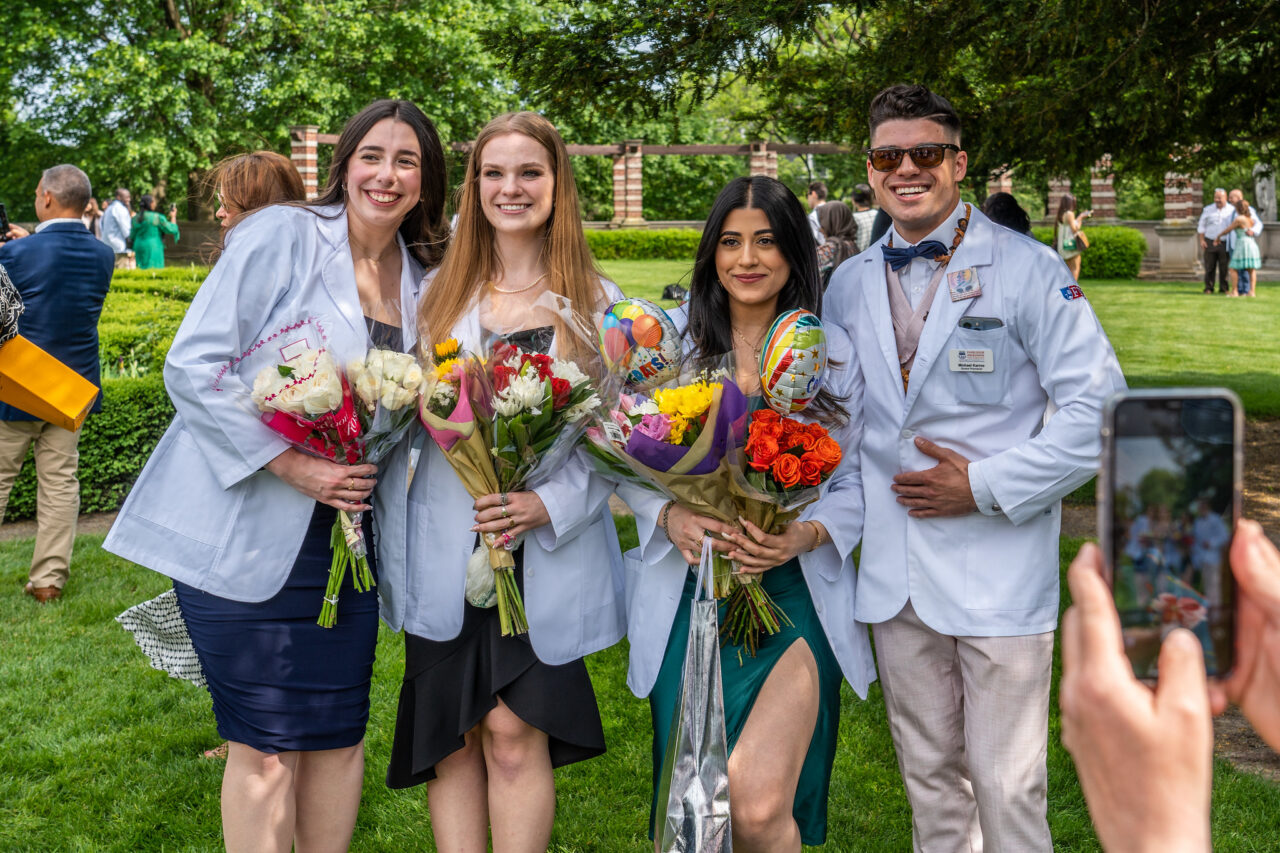 graduating students wear white coats and hold flowers.