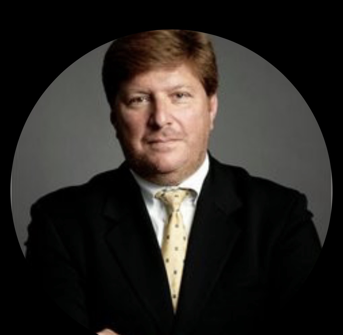 Headshot of Alan Chinich, a man with reddish-brown hair wearing a black suit jacket and a yellow patterned tie against a dark background.