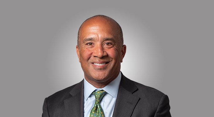Headshot of Clinton Lewis, a smiling bald man wearing a dark gray suit, light blue shirt, and a green patterned tie.