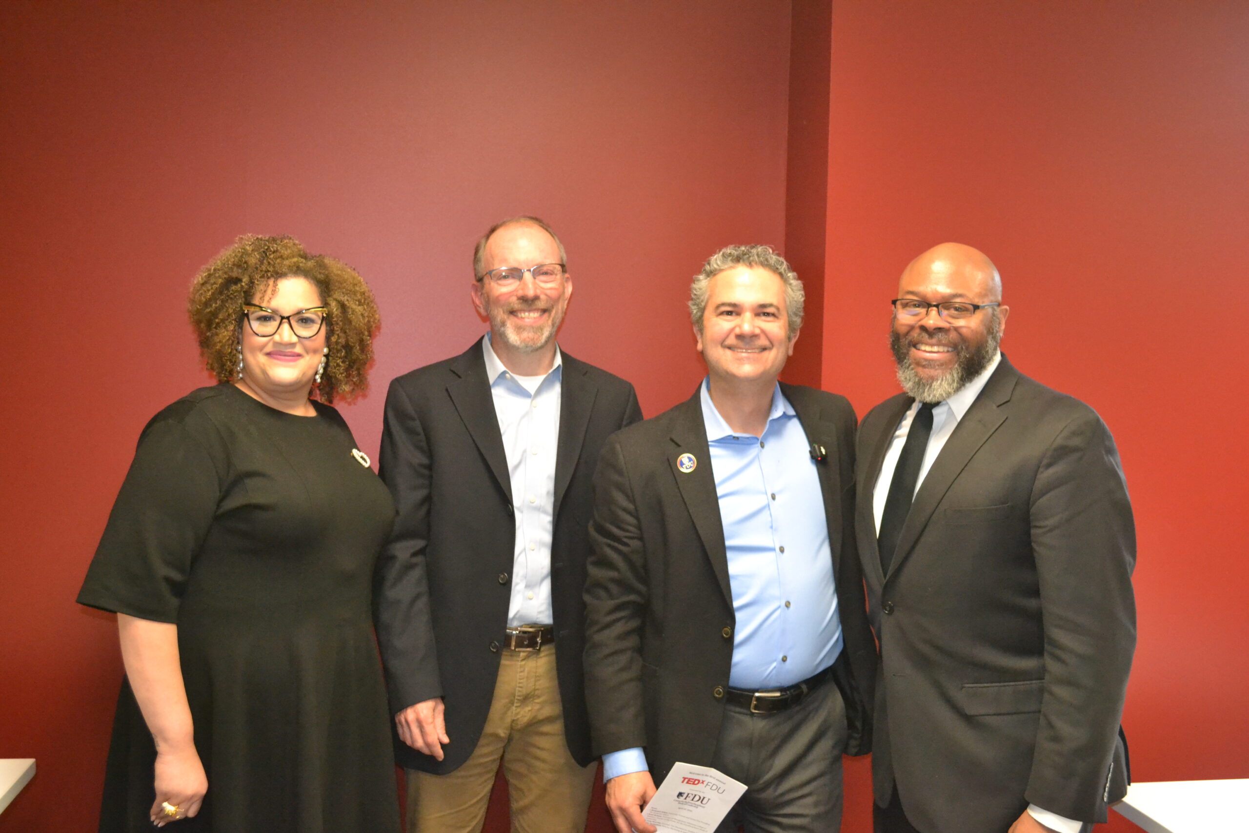 Four people stand in a row in front of a red wall.