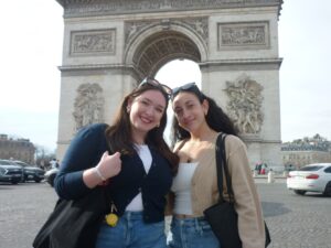 Two women smile in front of the Arc de Triomphe. Both wear cardigans over white tops with jeans and have sunglasses on their heads. The massive stone monument with sculpted reliefs stands behind them on a cobblestone plaza with cars visible.
