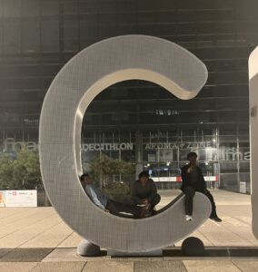 Three people pose with a large, grey, perforated metal sculpture shaped like a wide loop. One person stands behind it while two sit inside the curve. Behind them is a glass building with "DECATHLON" and "AROMA ZONE" text visible.