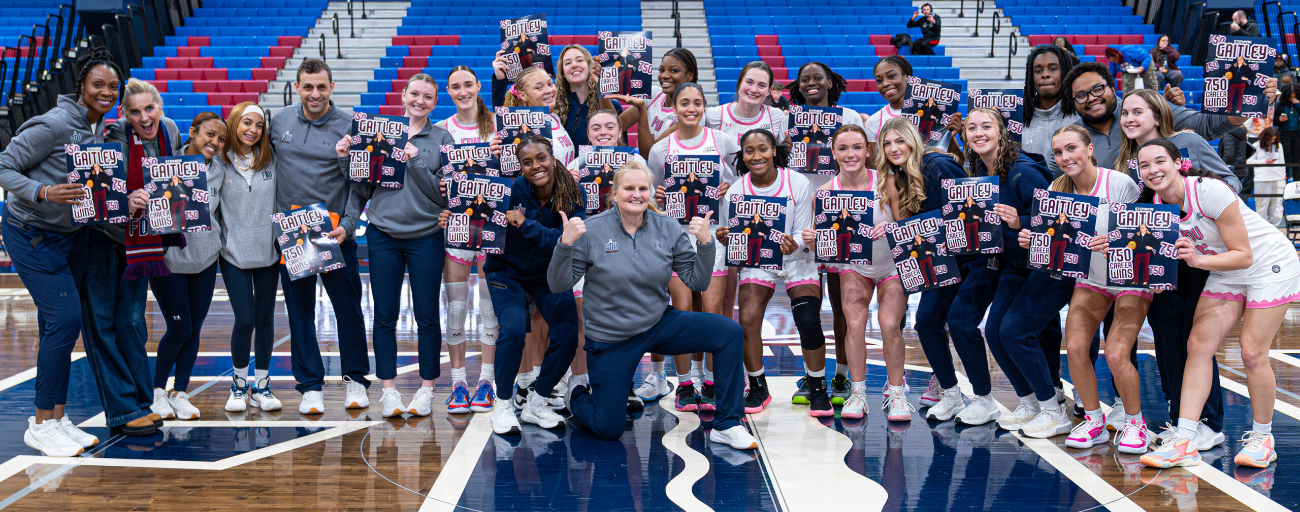 A female coach sits in front of her women's basketball team. Her players hold posters with their coach's face and the words 750 career wins.