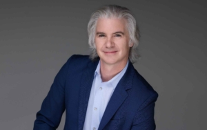 a man leans towards the camera and smiles. he wears a blue blazer.