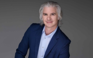 a man leans towards the camera and smiles. he wears a blue blazer.