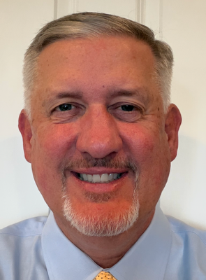 A headshot of Joseph Chesakis, a smiling man with short gray hair and a goatee, wearing a light blue collared shirt.