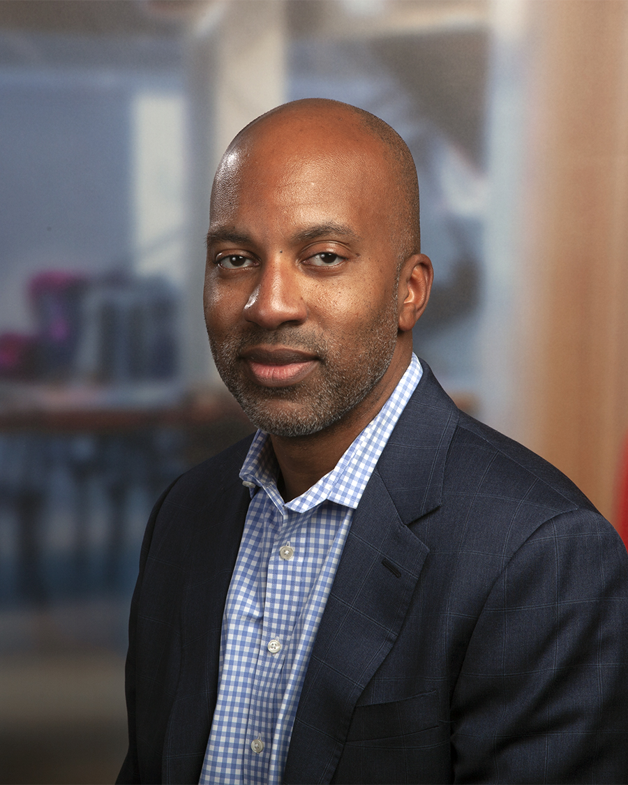 A headshot of Kyle Bolden, a man with a shaved head and goatee, wearing a dark blazer over a light blue patterned shirt.