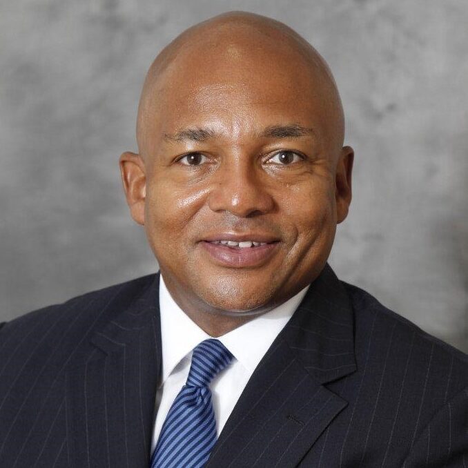 A headshot of Lester Owens, a smiling man with a shaved head, wearing a dark suit, white shirt, and a blue patterned tie.