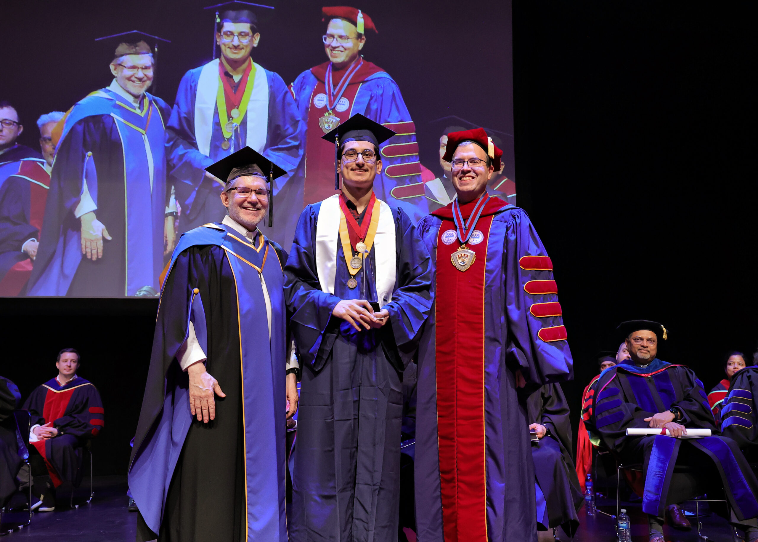 a graduating student stands on a stage with university leadership members. he holds an award.