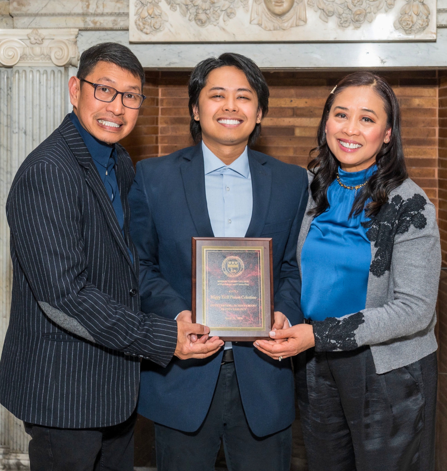 a college student and his parents smile at the camera. they hold a plaque award.