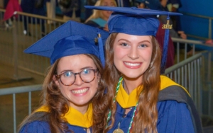 two graduating students smile at the camera. they wear caps and gowns regalia.