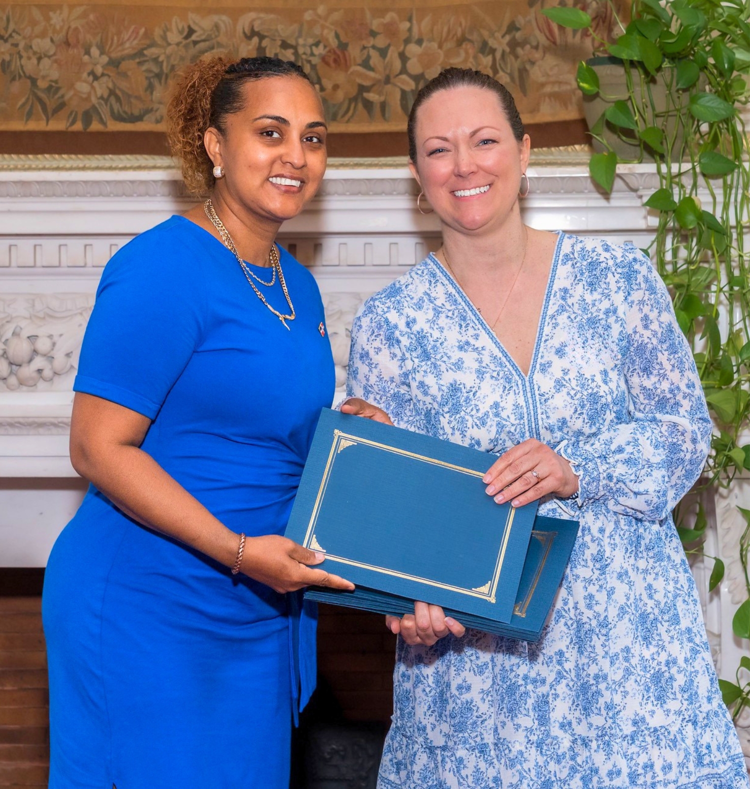 two women wear blue dresses and smile for the camera. they hold up a certificate folder.