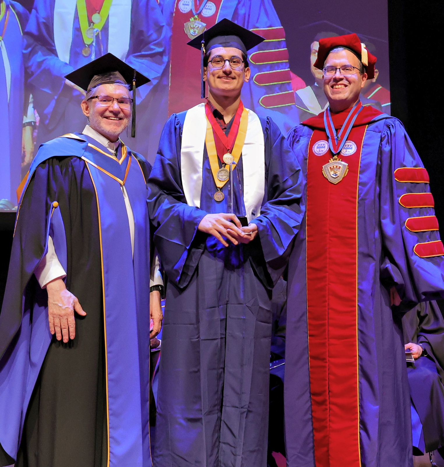 a graduating student stands on a stage with university leadership members. he holds an award.