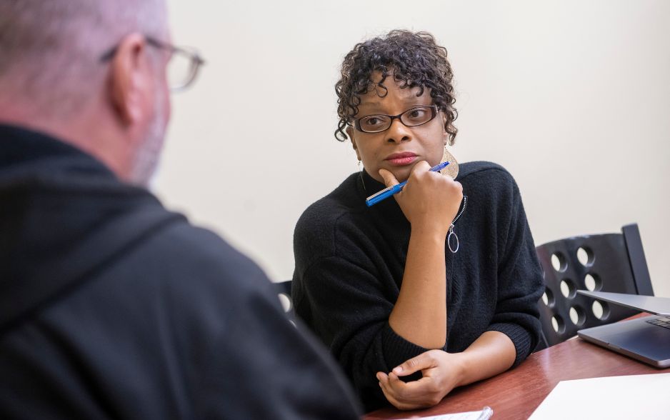 an adult learner listens to another person. she holds a pen and rests her head on her hand, as if she is thinking and concentrating.