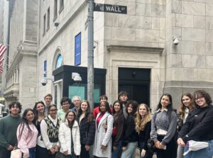 A group of young people stands on a cobblestone street corner in front of a stone building. A street sign reading "WALL ST" is visible on the building corner above them. An American flag hangs on the left side, and the group wears casual jackets and sweaters.