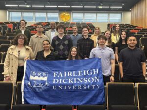 A group of students and faculty poses in a modern lecture hall with tiered seating and black chairs with orange accents. They hold a blue Fairleigh Dickinson University banner with the school's crest. Behind them, windows line the upper wall and a golden spherical sculpture is mounted between them.