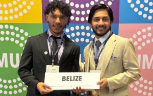 Two smiling students wearing students holding a card that says Belize in front of a multicolor NMUN backdrop.