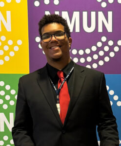 A smiling student wearing a black suit, red tie and glasses in front of a multicolor NMUN backdrop.