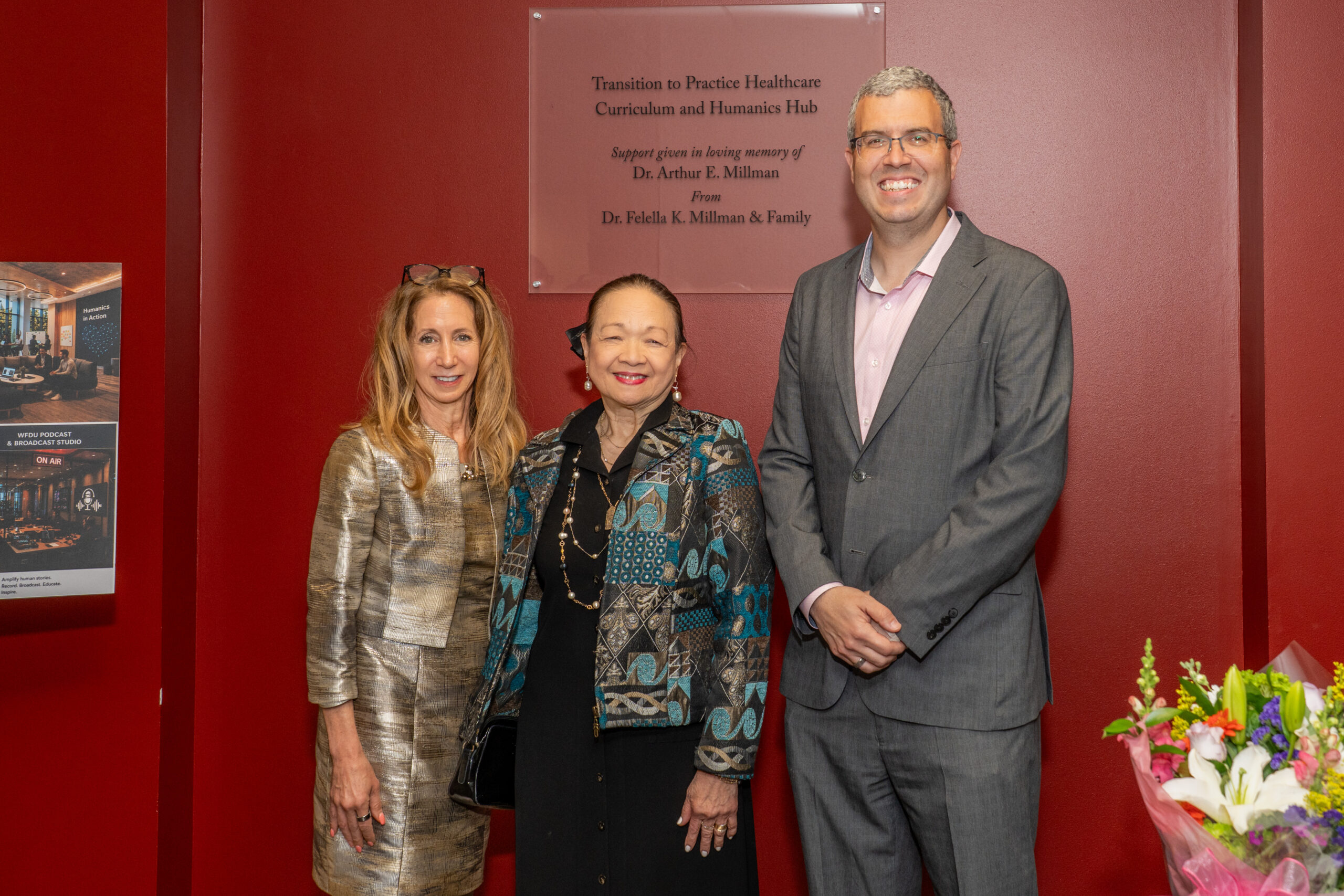 Three people stand together as a group in front of a plaque that is clear and a burgundy wall.