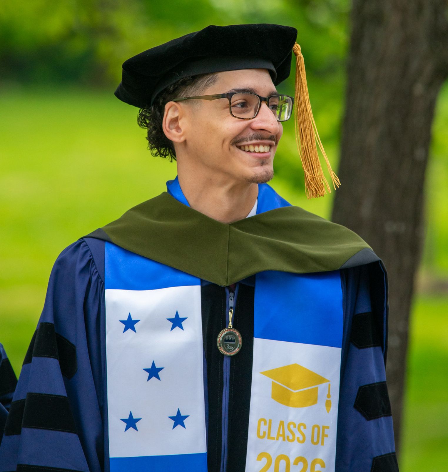 a doctoral student graduates. he wears academic regalia. he smiles at someone out of frame. he is outside.