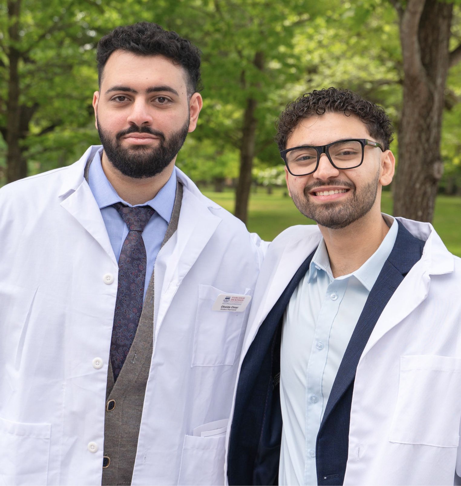 two students wear white coats and smile at the camera. they are outside.