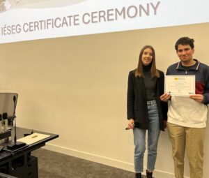 A young man and woman stand on a stage in front of a large projection screen displaying "IESEG CERTIFICATE CEREMONY". The woman holds a microphone and wears a black blazer, while the man holds a participation certificate and wears a color-blocked sweater. They are dressed in smart-casual attire.