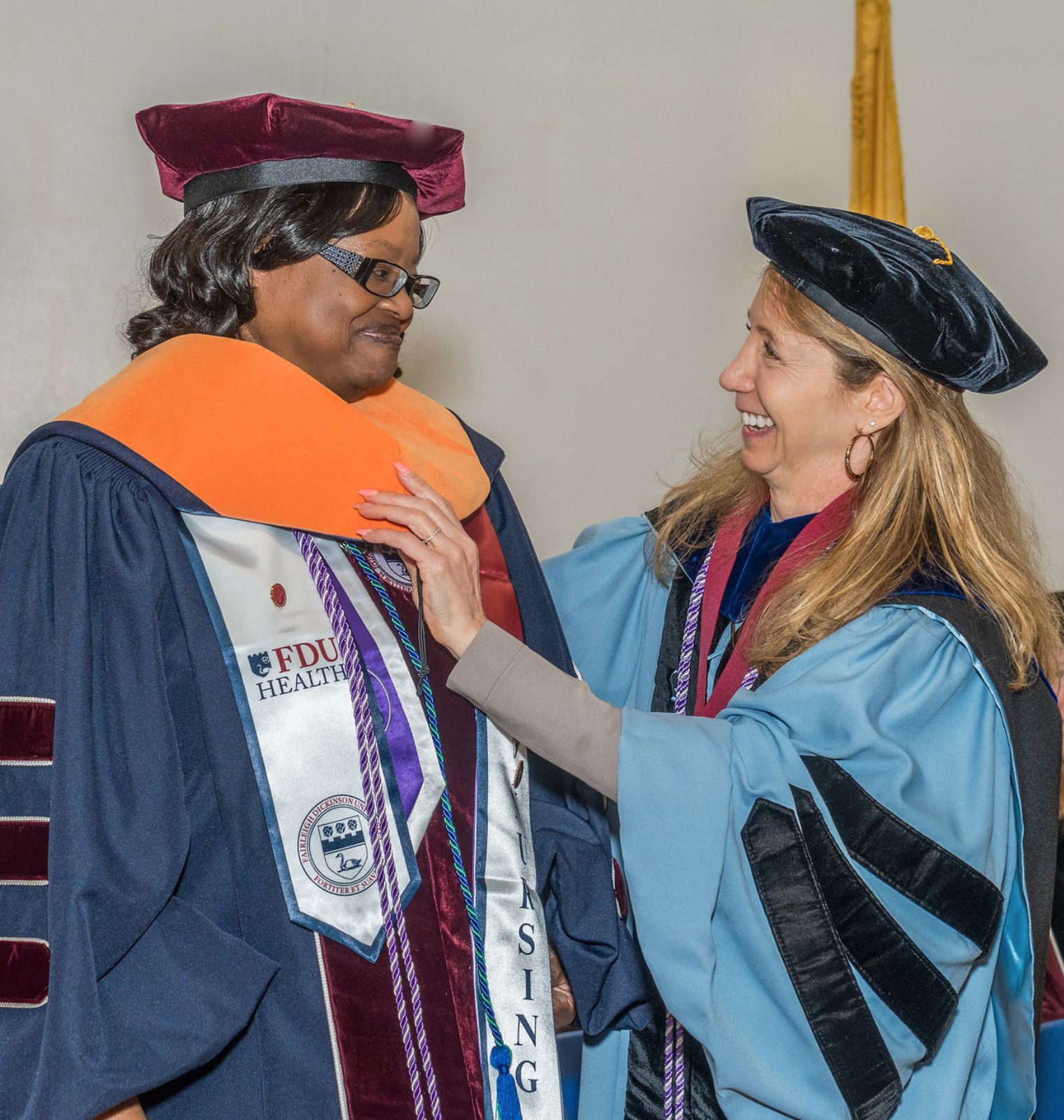 a doctoral student graduates. she wears academic regalia. a professor adjusts her hood and they smile at each other.