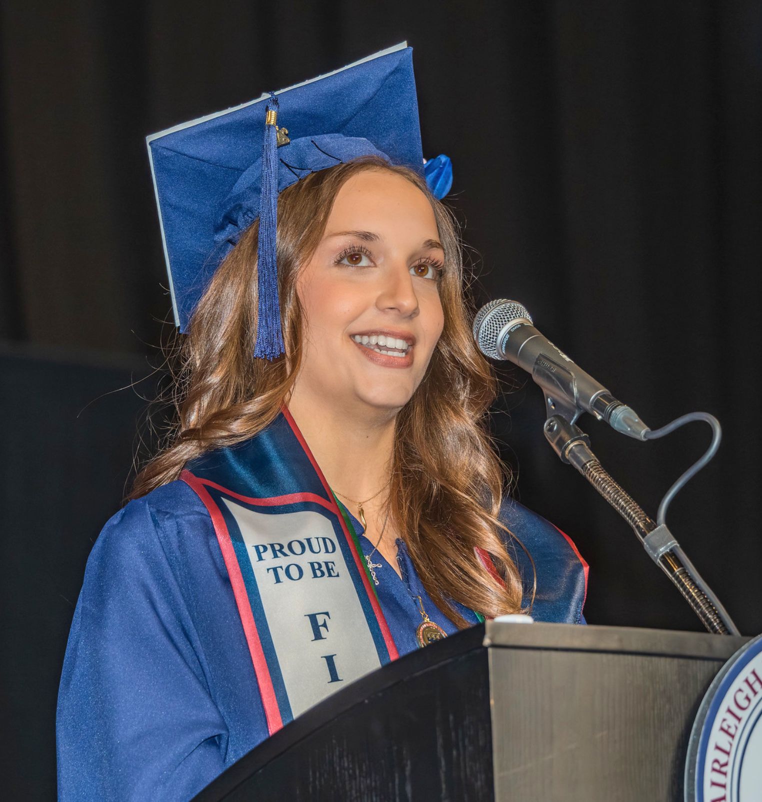 a graduating student speaks into a microphone at a podium. she wears a cap, a gown and various cords and stoles.