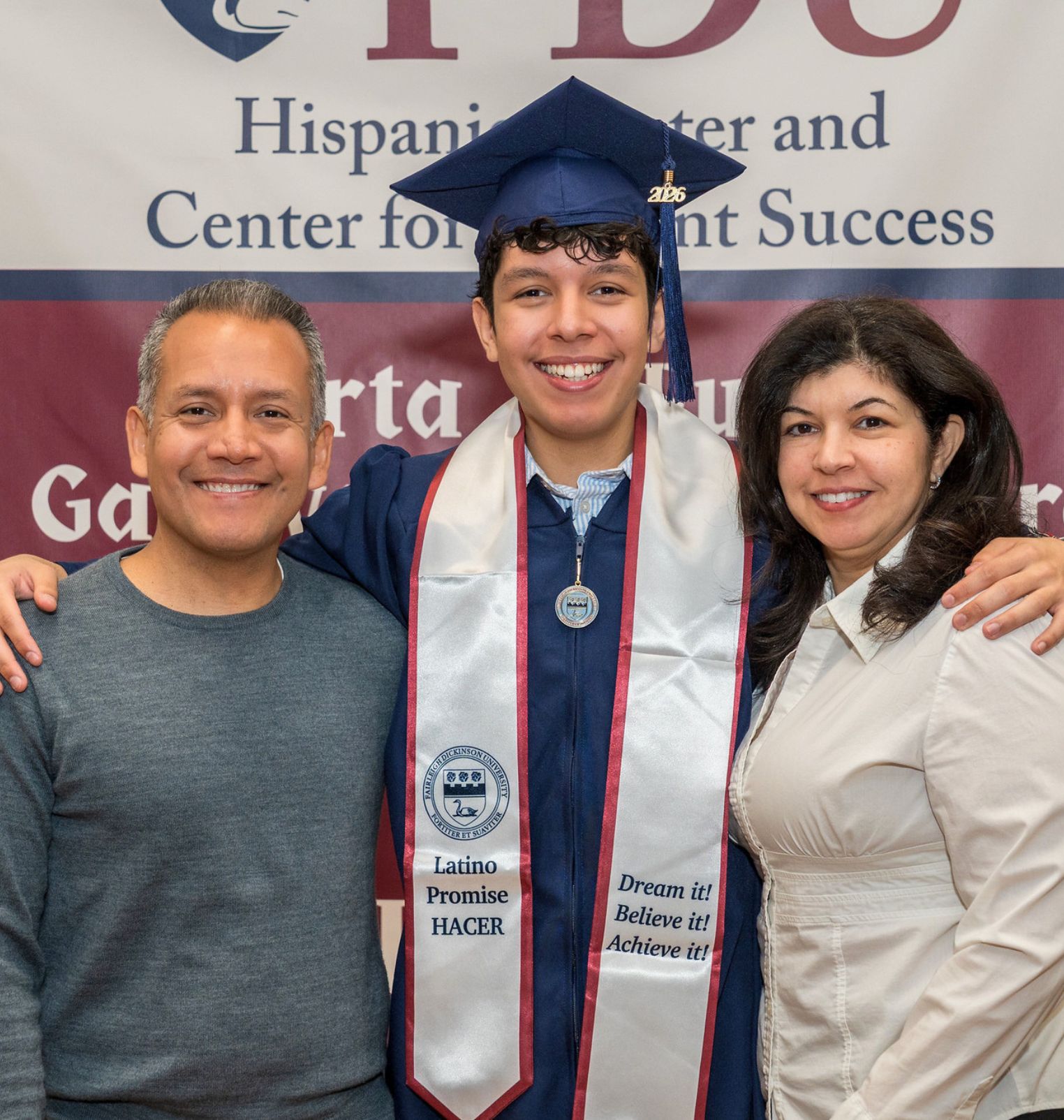 a college student and his parents smile at the camera. the student wears a cap and gown. behind them reads a sign 