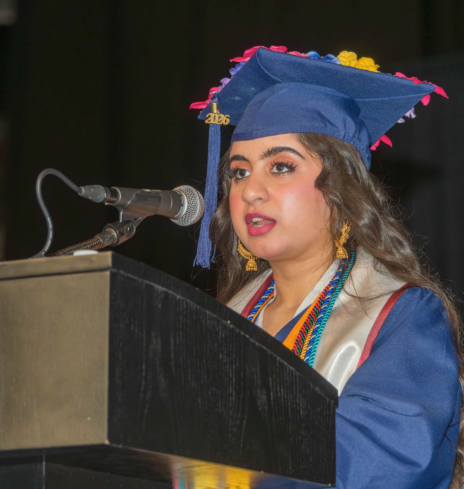a graduating student speaks into a microphone at a podium. she wears a cap, a gown and various cords and stoles.