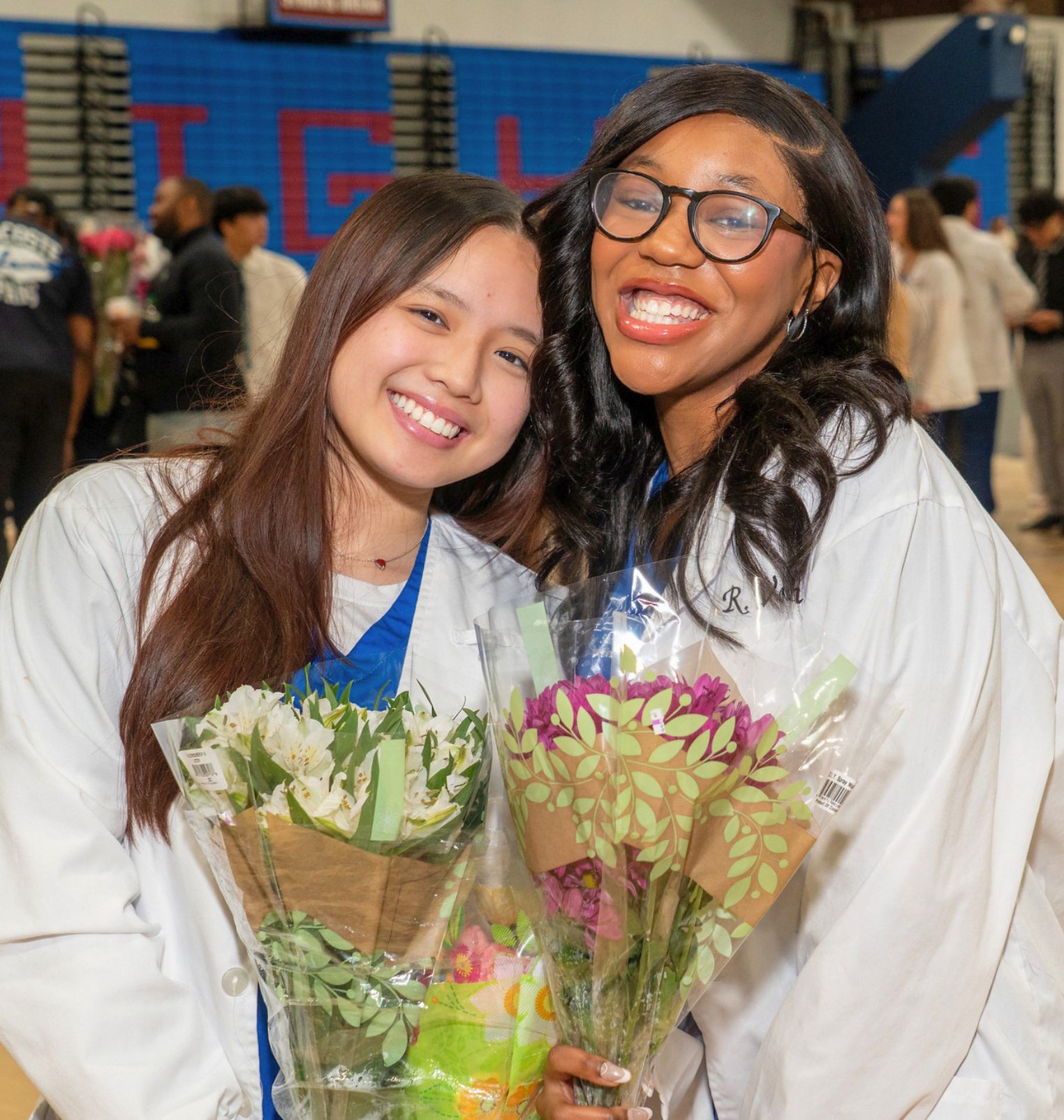 two students wear white coats and hold flowers. they lean together and smile.