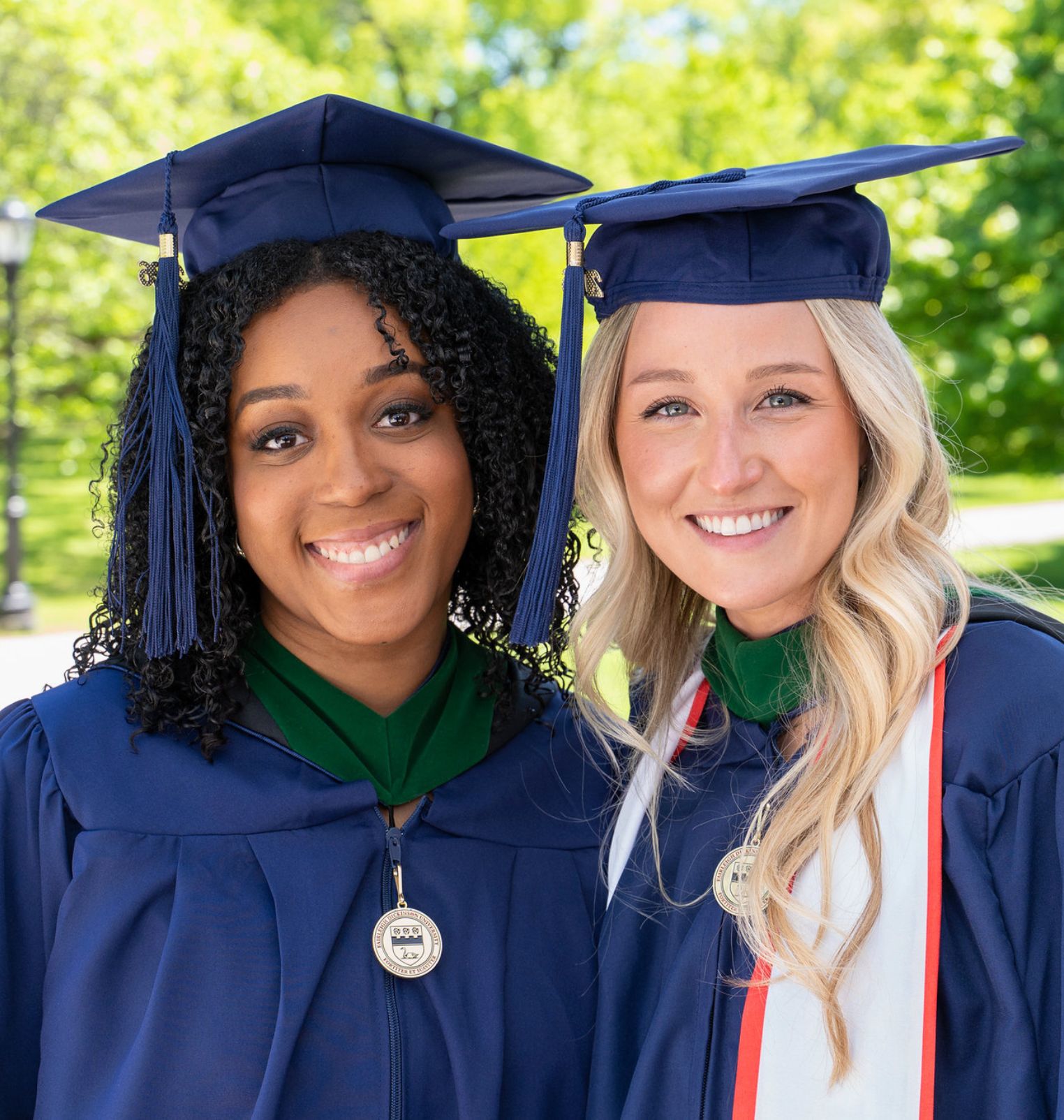two graduating students stand outside and smile. they both wear caps, gowns and hoods.