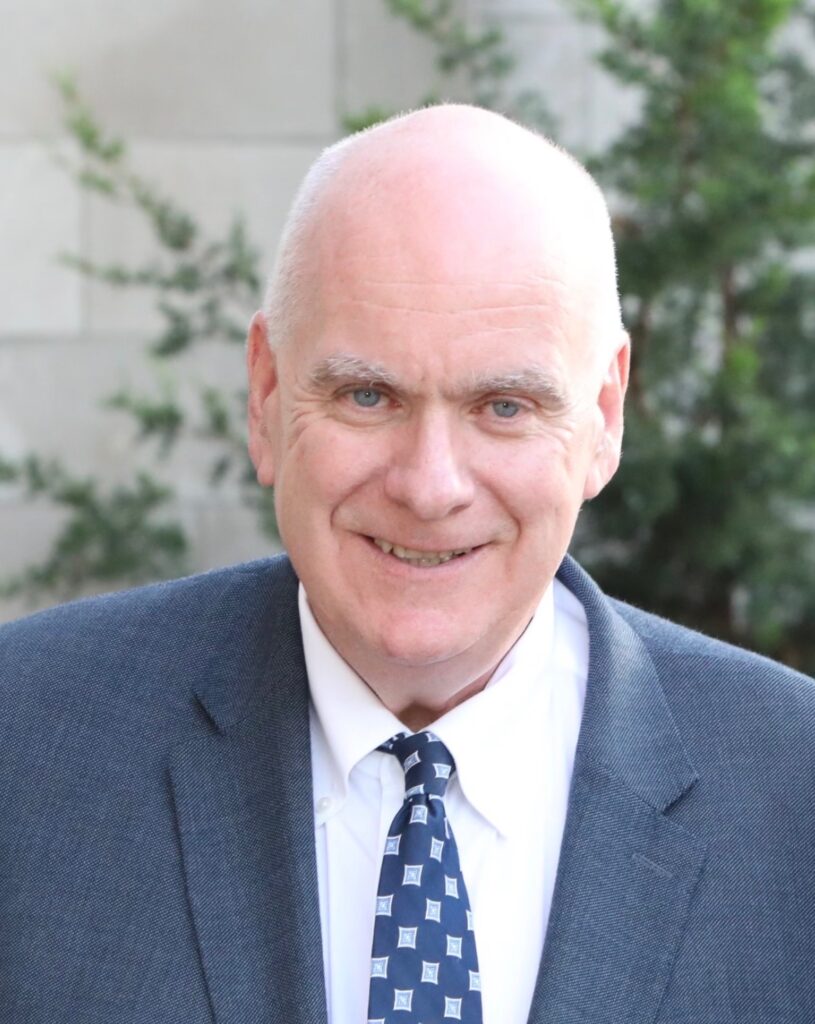 Professional headshot of David Conroy, a smiling man with short gray hair on the sides and blue eyes. He is wearing a textured blue-gray suit jacket, a white dress shirt, and a navy blue tie with a light blue geometric pattern, photographed outdoors against a blurred background of green foliage and a light-colored wall.