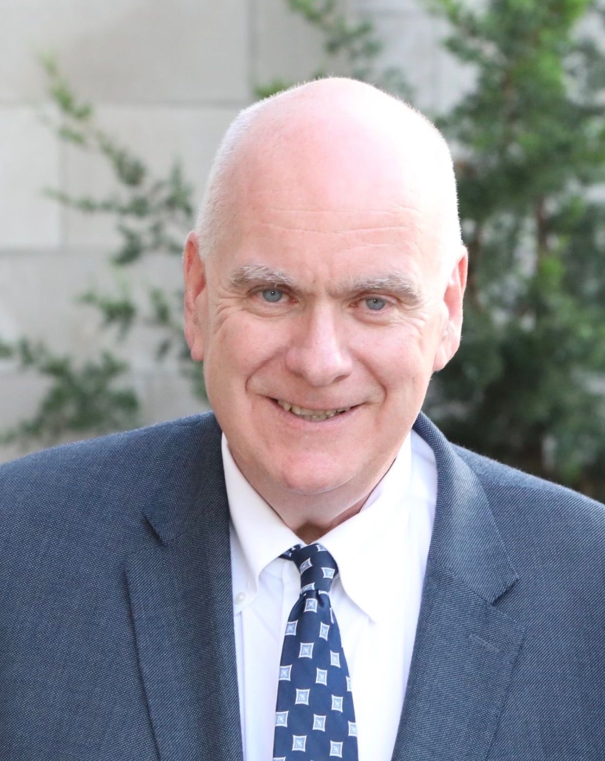 Professional headshot of David Conroy, a smiling man with short gray hair on the sides and blue eyes. He is wearing a textured blue-gray suit jacket, a white dress shirt, and a navy blue tie with a light blue geometric pattern, photographed outdoors against a blurred background of green foliage and a light-colored wall.