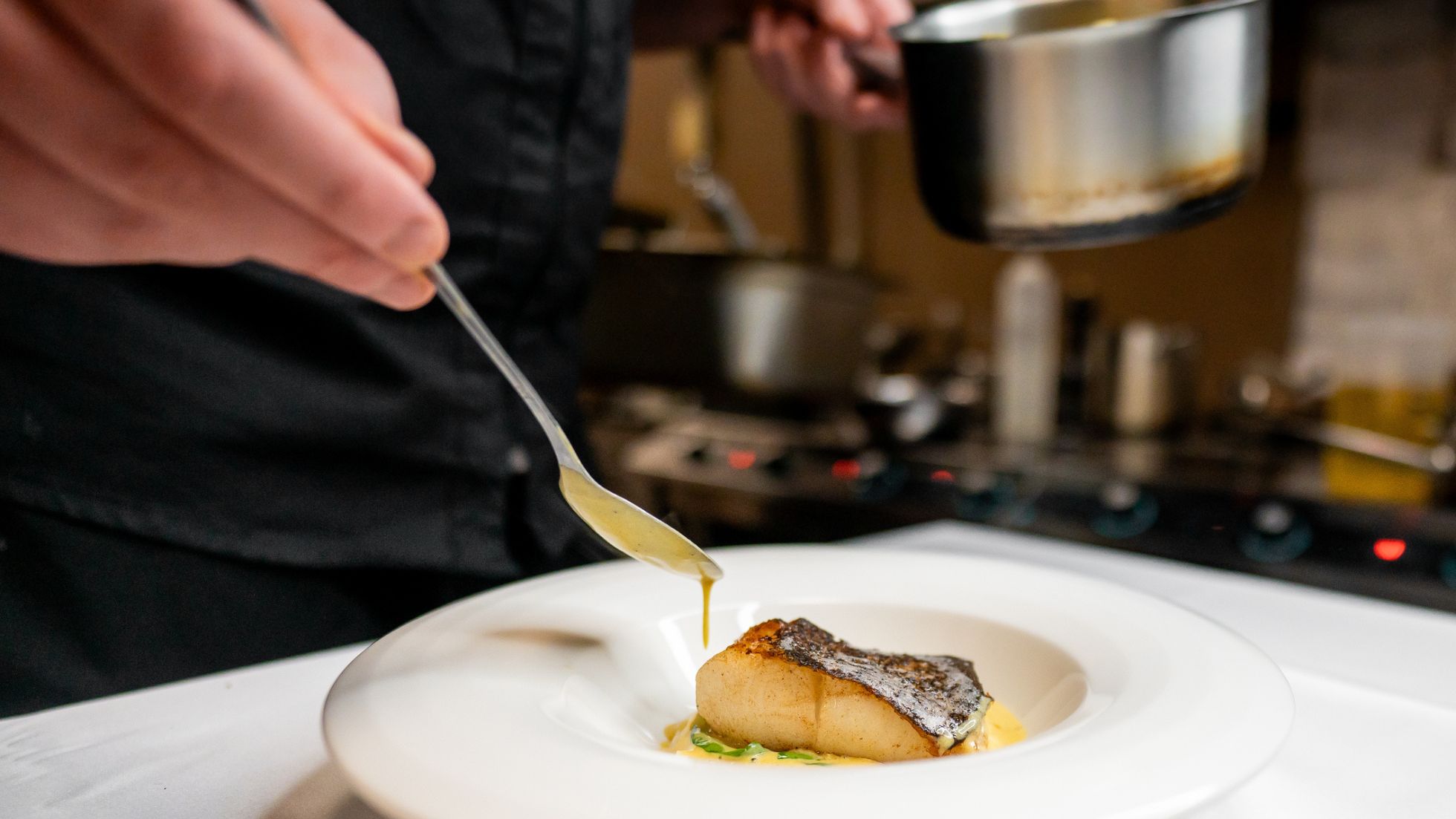 a chef plates a fine dining meal. he drizzles a sauce onto fish. the photo is a close-up of the chef's hands and the plate.