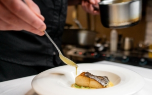 a chef plates a fine dining meal. he drizzles a sauce onto fish. the photo is a close-up of the chef's hands and the plate.