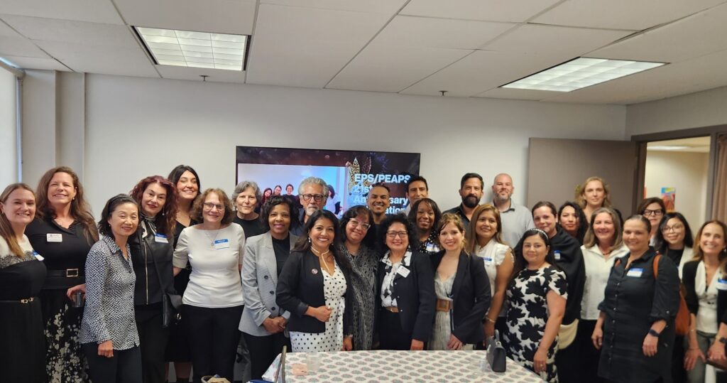 A group photo of 28 people at the EPS/PEAPS anniversary celebration, taken in an office conference room with fluorescent lighting. Attendees are dressed in business casual attire and wearing name tags, gathered in front of a banner/screen displaying the event information.
