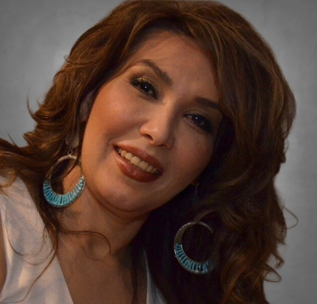 A professional headshot of a woman with wavy brown hair wearing large turquoise statement hoop earrings. She is wearing a white or light-colored top and smiling warmly at the camera against a neutral gray background.