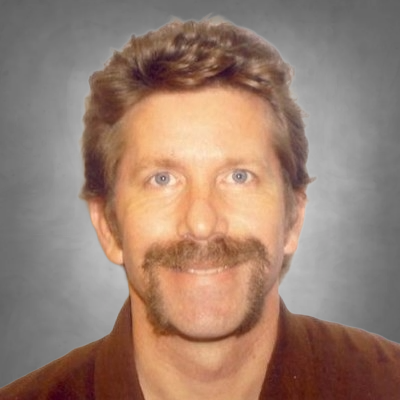 A professional headshot of a man with short light brown hair and blue eyes. He has a mustache and a small goatee. He is wearing a brown collared shirt and has a slight smile, photographed against a neutral gray background.