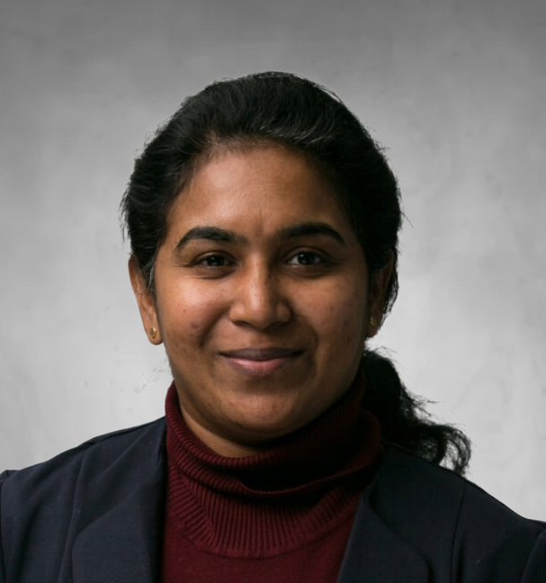 A professional headshot of a woman with dark hair pulled back. She is wearing a maroon turtleneck sweater under a dark blazer or jacket. She has a gentle, pleasant smile and is photographed against a neutral gray background.