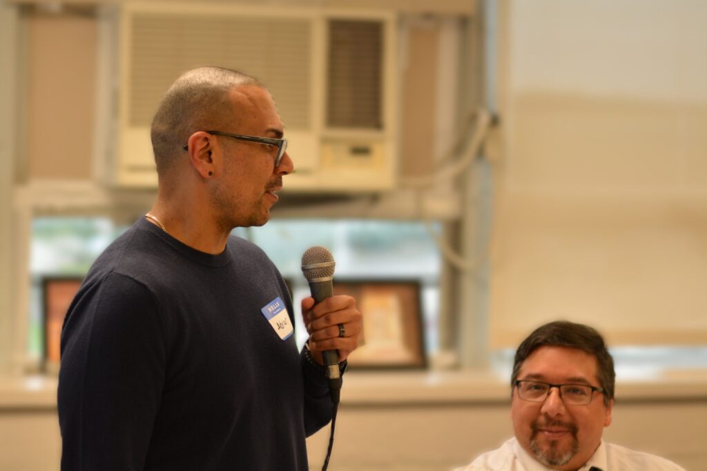 A man with glasses and a shaved head, wearing a dark blue shirt with a name tag, speaks into a microphone at an indoor event. Another attendee is partially visible in the background.